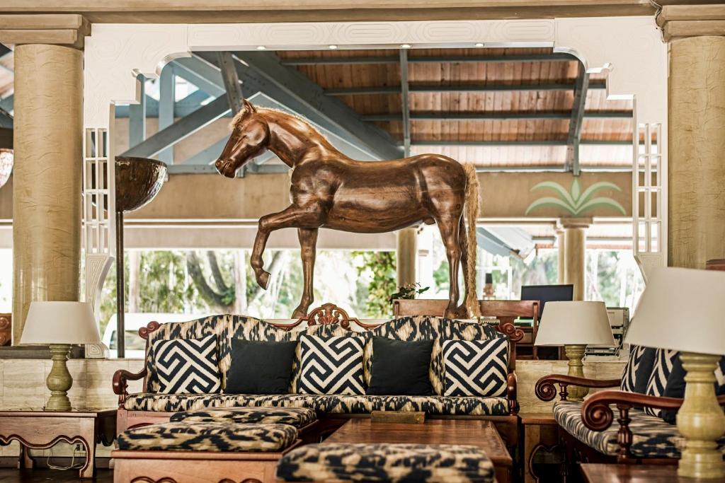 Lieliska viesnīcas interjera ainava ar koka zirdziņu skulptūru, kas novietota virs eleganta dīvāna ar ģeometriskiem spilveniem. Interjers ir stilīgs un aicinošs, radot mājīgu atmosfēru.