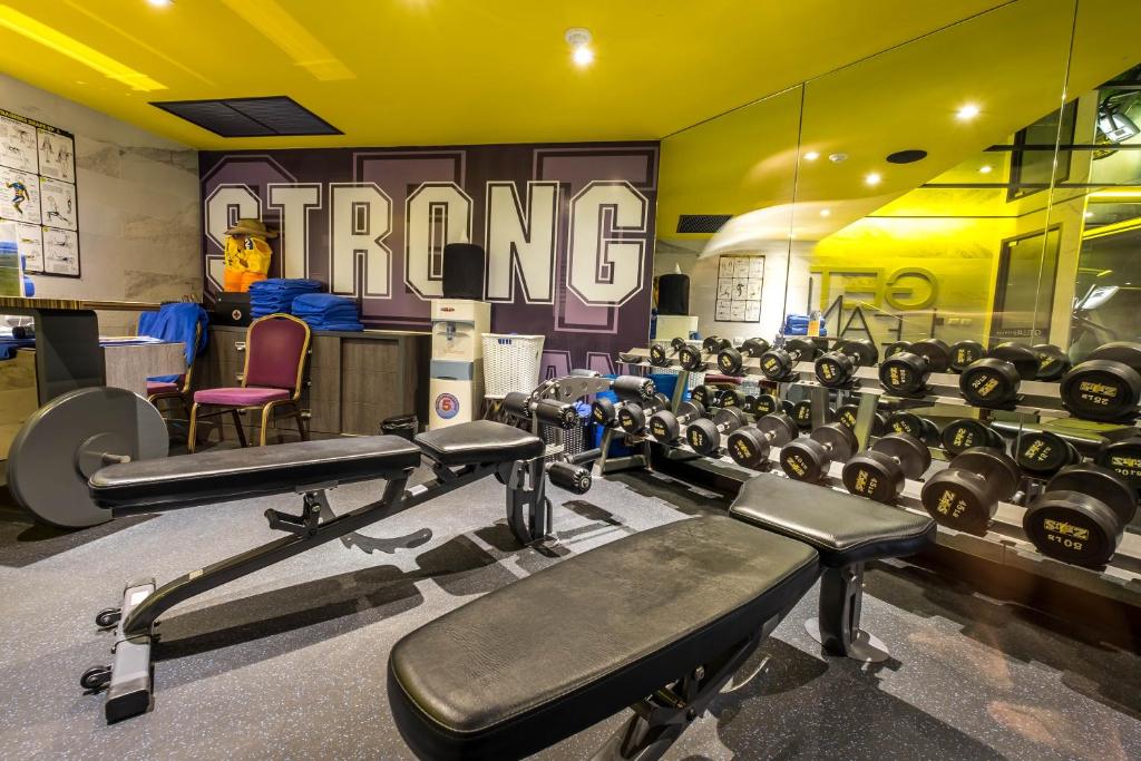 Mūsdienīga sporta zāle ar dzelzs svaru komplektu un treniņu soli, uzsverot "STRONG" tematu uz sienas. Telpā valda spilgts dzeltenais apgaismojums, radot motivējošu atmosfēru treniņiem.