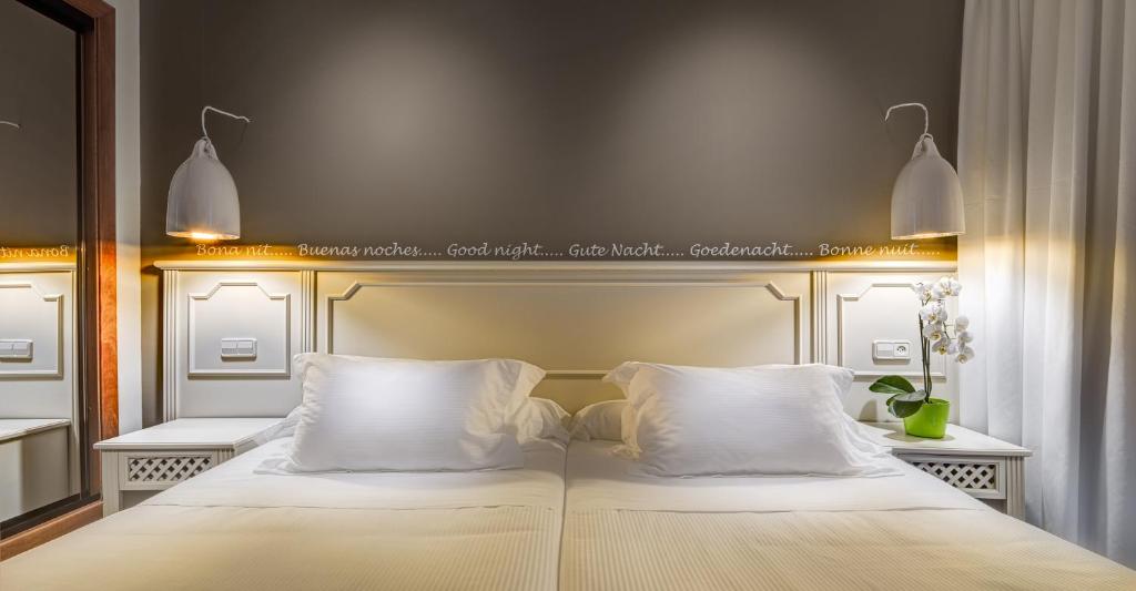 Mūsdienīgs guļamistabas interjers ar divām gultām, apgaismojumu un noformējumu, kas ietver dažādas valodas, kas sveic "labo nakti". Eleganta auga klātbūtne papildina atmosfēru.