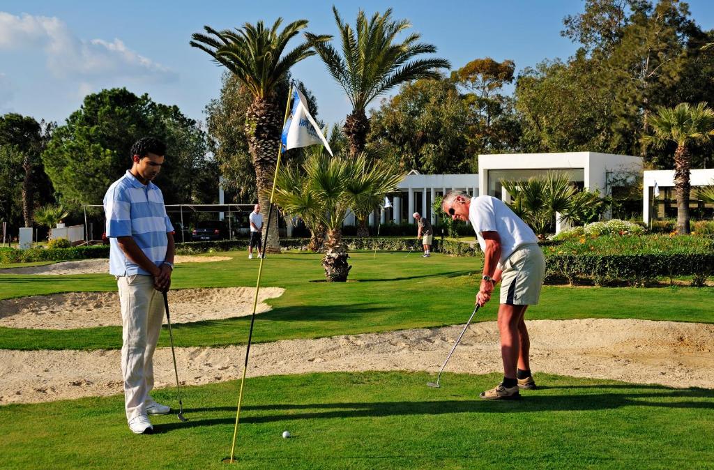 Golfspēles ainava ar diviem spēlētājiem, no kuriem viens sagatavojas sitienam pie bumbas, bet otrs gaida blakus. Apkārt ir zaļš zāliena laukums ar palmu kokiem un modernu ēku fona.