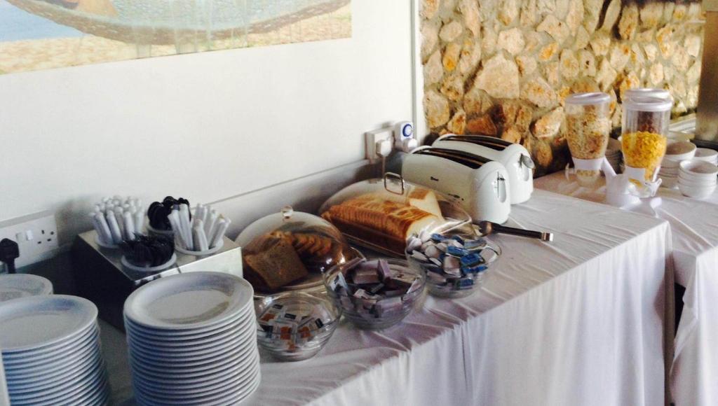 Brokastīm sagatavots bufetes galds ar šķīvjiem, maizes tosteri, cepumiem un graudaugu traukiem. Galds ir dekorēts ar baltu galdautu un atrodas pie akmens sienas.