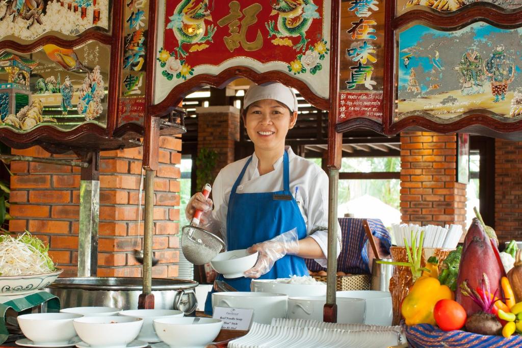 Sieviete, tērpta šefpavāra priekšautā, pasniedz ēdienu tirgus stendā. Fons izrotāts ar krāsainiem attēliem un ēdieniem, bet priekšplānā redzami svaigi dārzeņi un augļi.