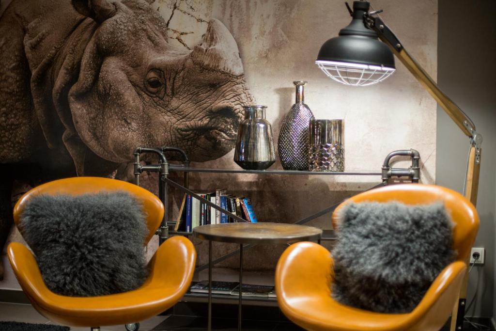 Mūsdienīgs interjers ar divām oranžām krēslām un pelēku pūkainu spilvenu, pie dzelzs galda ar stilīgām dekorācijām un lampu, pretī rhinoceros sienas gleznojuma.