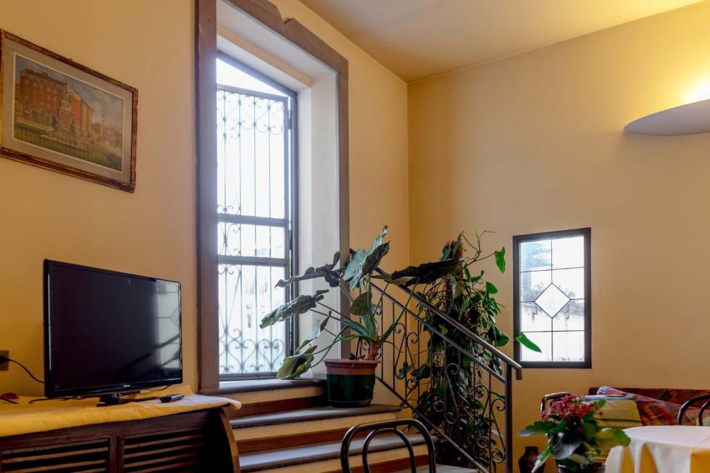 Mājas iekšējā telpa ar kāpņu telpu, televizoru, augu dekorācijām un logiem, kas sniedz gaismu un skatu uz ārpusi.