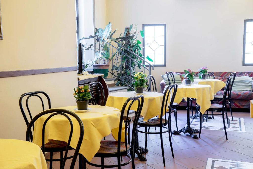 Kafejnīcas interjers ar dzeltenām galdautām un ziediem uz galdiem, gaišas sienas un daudz dabīgas gaismas. Šeit ir redzams arī dīvāns un augi, kas piešķir mājīgu atmosfēru.