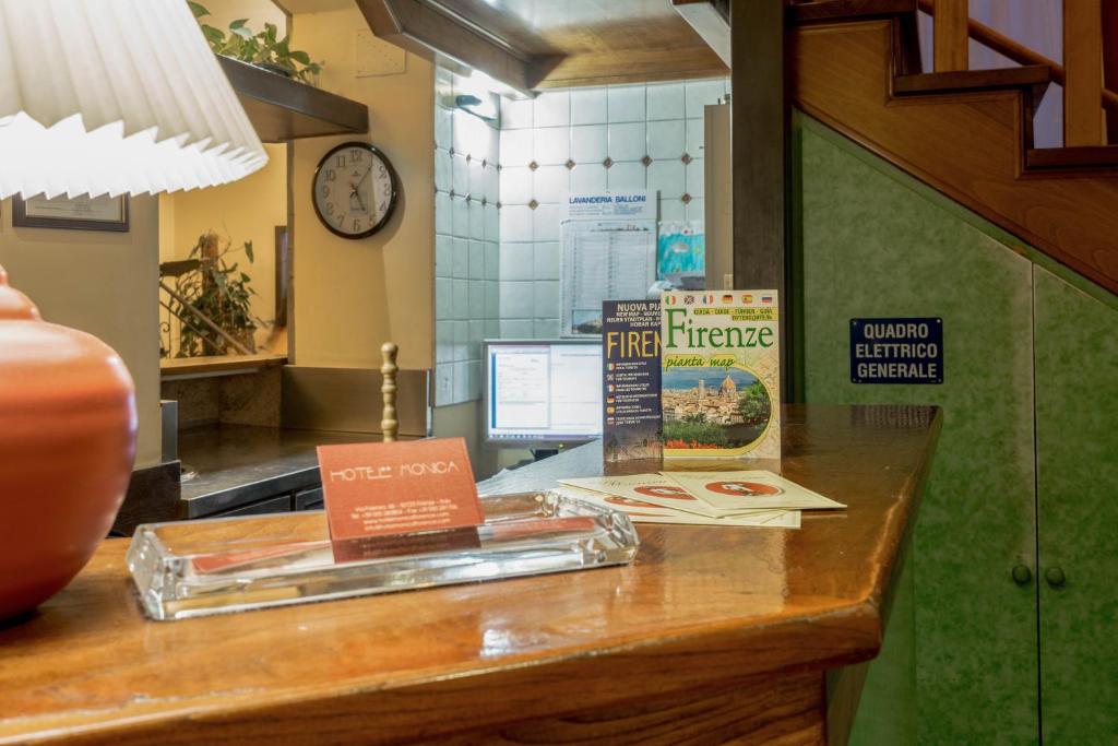 Reģistratūras zona viesnīcā ar koka galdu, lampu un informācijas materiāliem par Florenci. Redzama pulksteņa un datora ekrāns, kas norāda uz viesnīcas pakalpojumiem.