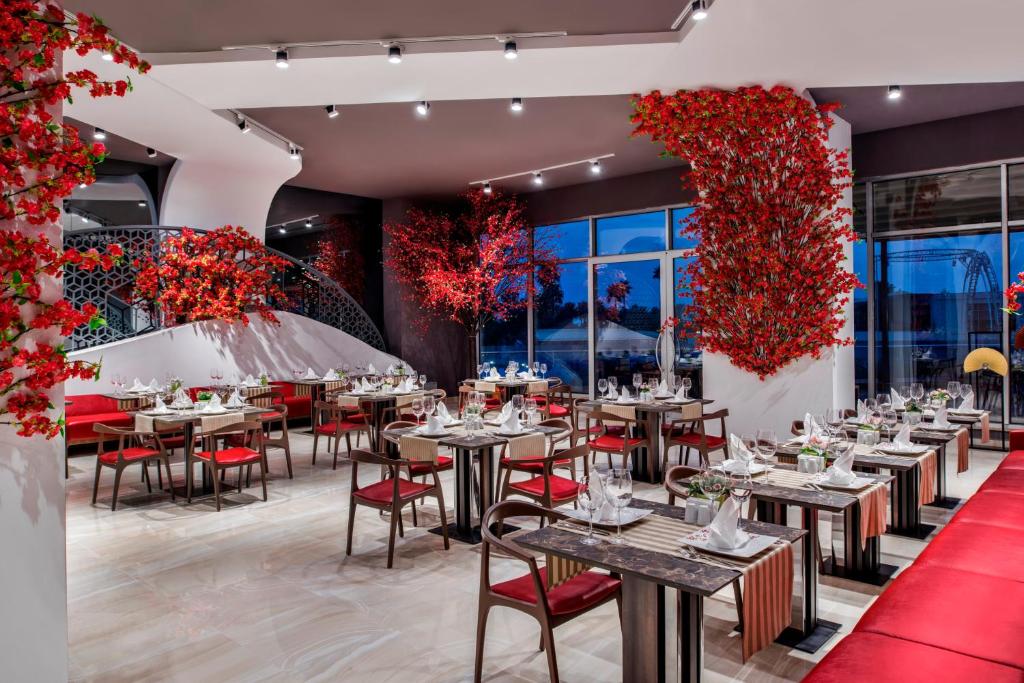 Mūsdienīgs restorāns ar elegantu interjeru, kurā ir sarkanas dekoratīvas augu kompozīcijas un stilīgi galdi. Gaismas apgaismojums un sarkanā krāsa rada siltu un patīkamu atmosfēru.