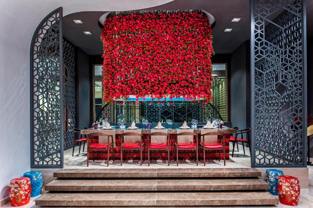 Mūsdienīgs restorāna interjers ar elegantu galdu un spilgtiem sarkaniem ziediem uz sienas. Dekorācijas ietver izsmalcinātus krēslus un krāsainus sēdvietas, radot patīkamu atmosfēru.