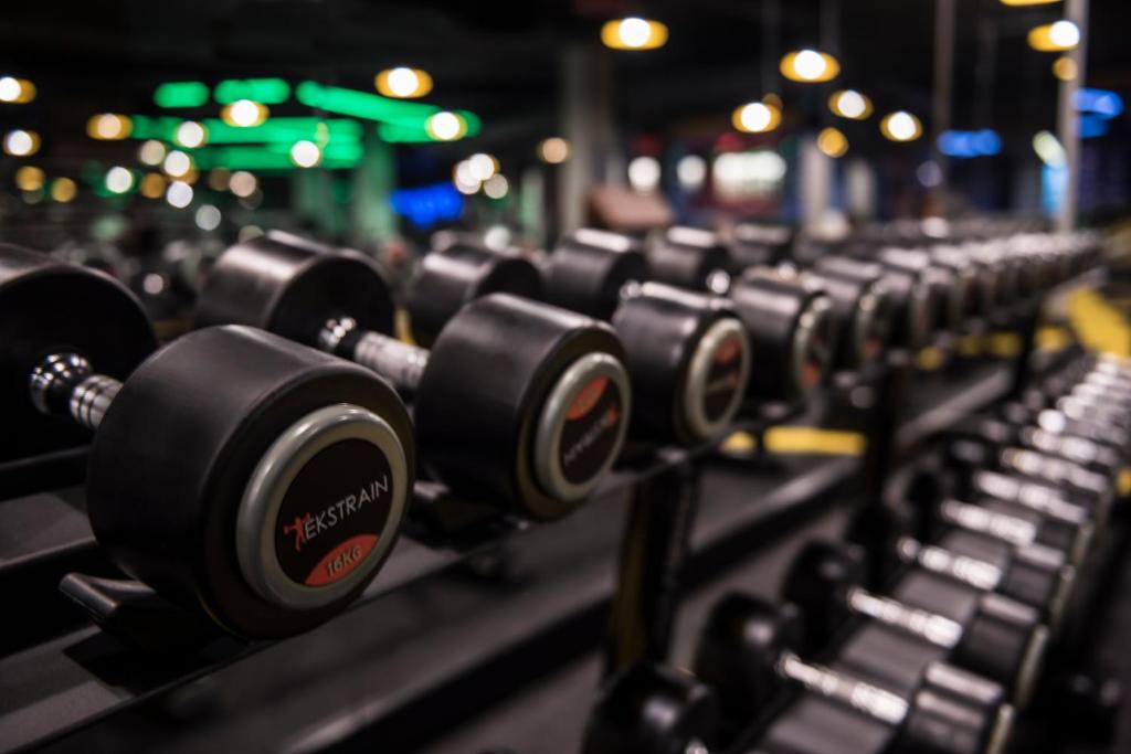 Svaru rinda sporta zālē ar dažādiem smagumiem, uzsverot fitnesa un treniņu vidi. Apgaismojums rada dinamisku atmosfēru.