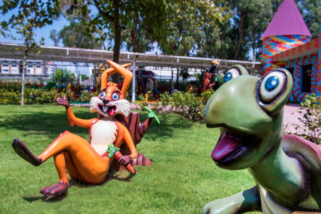 Krāsaini un jautri dzīvnieku tēli, kas attēlo zaļu bruņurupuci un oranžu zaķi, atpūšas saulainā parkā. Fons ar košām dekorācijām un ziediem papildina skatu.