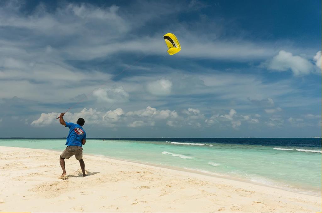 Vīrietis lidinās ar dzeltenu izpletni pludmalē zem zila debesu fona. Jūras viļņi mierīgi plūst krastā, radot idillisku skatu.