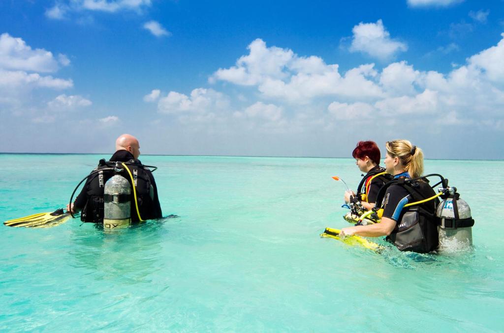 Trīs nirēji, tērpti niršanas ekipējumā, dodas ūdenī ar skaidru, zilu debesi fona. Viens no viņiem tur nirstošo dēli, bet citiem ir niršanas tvertne uz muguras.