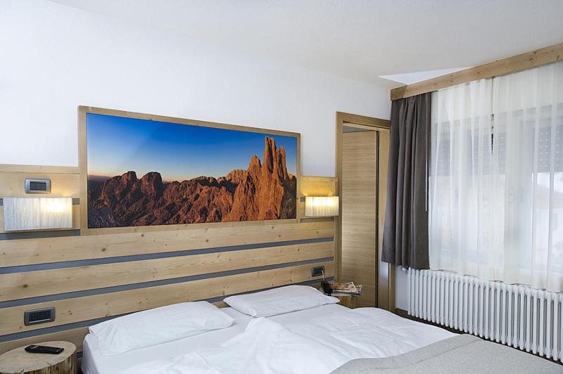 Mūsdienīgs guļamistabas interjers ar koka sienu apdari un gultu, virs kuras ir ainavas attēls. Telpā ir lieli logi ar caurspīdīgām aizkariem, radot gaišu un mājīgu atmosfēru.