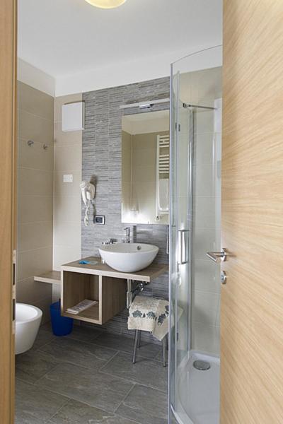 Mūsdienīga vannas istaba ar stikla dušu, pelēkiem flīzēm un elegantu izlietni uz koka plaukta. Interjers veidots ar minimālistisku pieeju, uzsverot funkcionalitāti un estētiku.