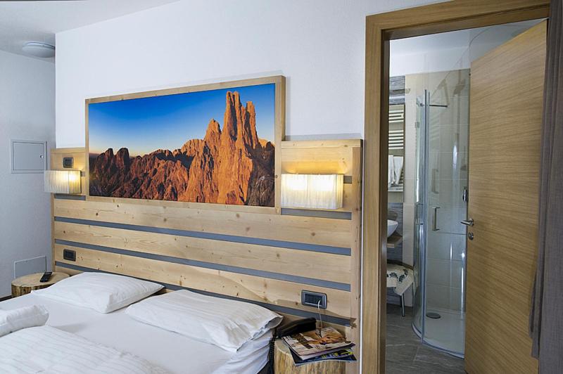 Mūsdienīga guļamistaba ar koka gultu un sienas gaismekļiem. Virs gultas ir ainavu attēls, kas papildina telpas noskaņu. Blakus ir vannas istaba ar stikla durvīm.