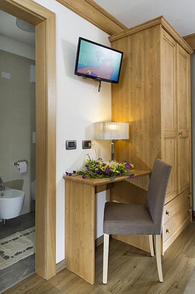 Mūsdienīga guļamistaba ar koka mēbelēm, rakstāmo un televizoru, kas piestiprināts pie sienas. Uz galda ir lampiņa un ziedu kompozīcija, radot mājīgu atmosfēru.