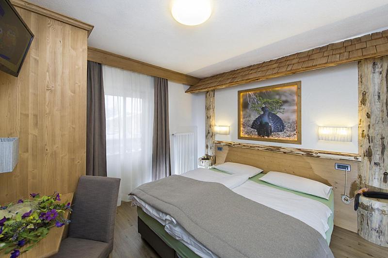 Mūsdienīgs guļamistabas interjers ar koka akcentiem, ērtu gultu un mākslas darbu uz sienas. Gaismas avoti rada siltu atmosfēru, savukārt logs nodrošina dabīgu apgaismojumu.