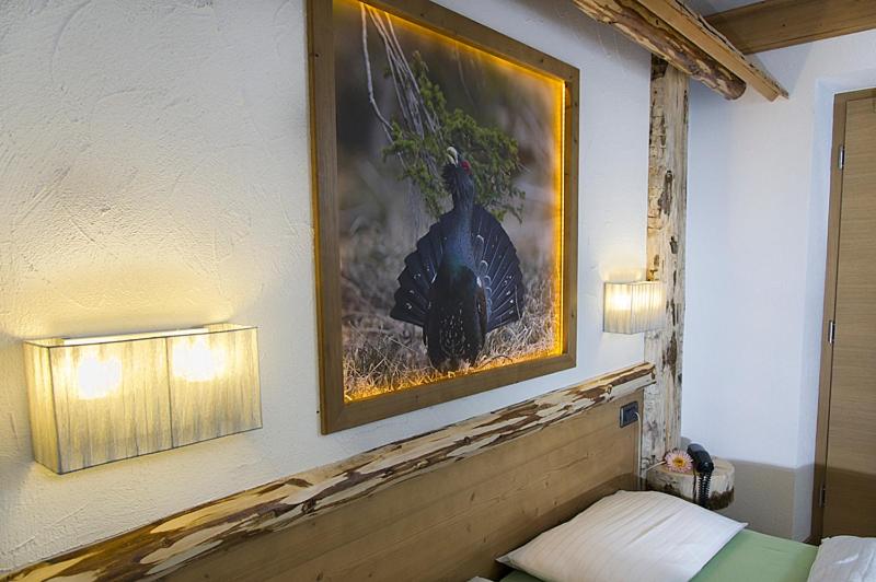 Mūsdienīgs viesnīcas numurs ar koka apdari, kurā redzama izsmalcināta bilde ar melno gārni, kas apgaismota ar LED gaismu. Sienas lampas pie gultas papildina telpas atmosfēru.