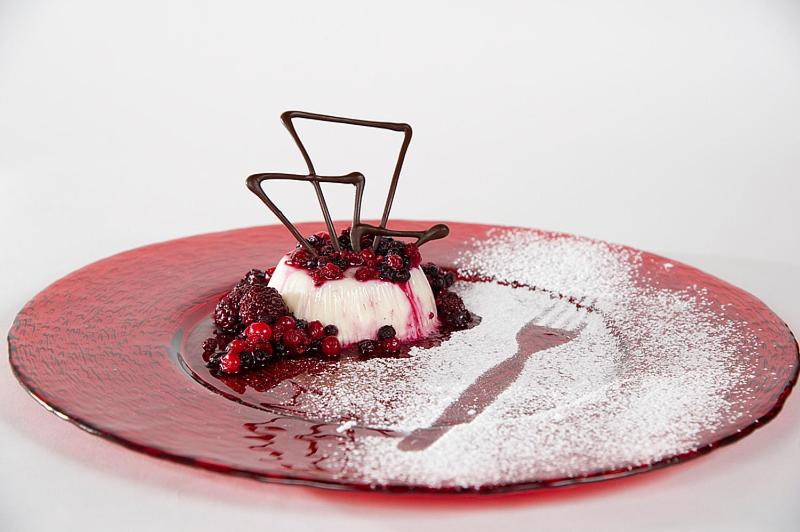 Dekorēta deserts uz sarkanas šķīvja ar ogu mērci un šokolādes dekorāciju. Izsmalcināts un estētiski pievilcīgs ēdiens.