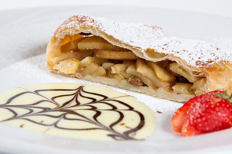 Ābolu strūdelis ar pūdercukuru, blakus krēmveida mērce un zemeņu šķēle.