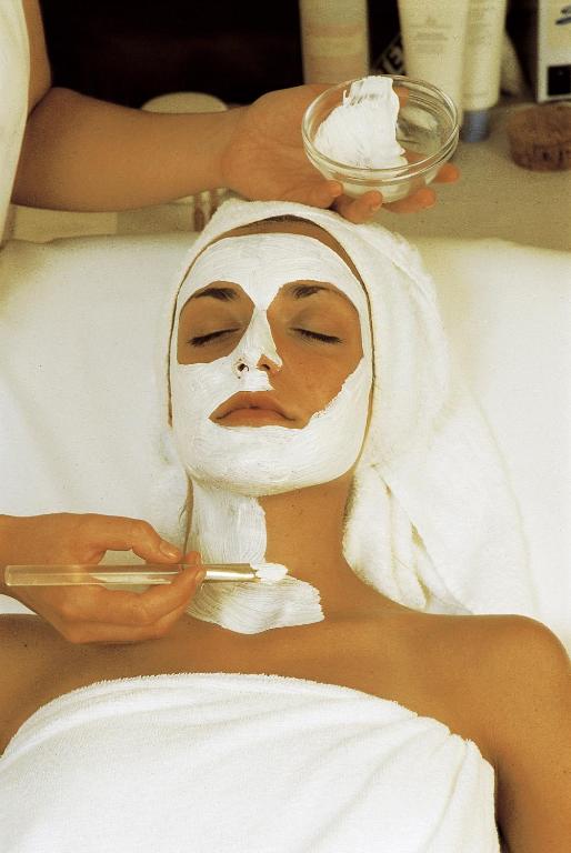 Sejas maskas uzklāšana spa procedūrā, sieviete ar baltu masku uz sejas un dvieli ap galvu. Mierīga un relaksējoša atmosfēra.