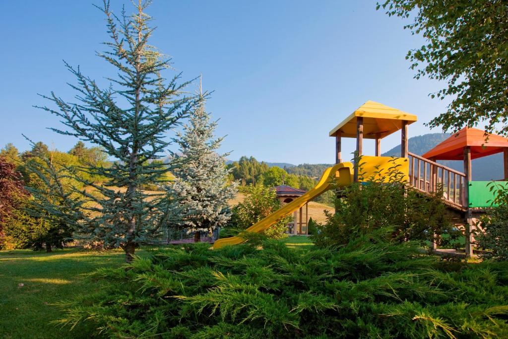 Bērnu rotaļu laukums ar dzeltenu slaidu un krāsainām nojumi, ko ieskauj zaļi krūmi un priedes, dabas vidē.