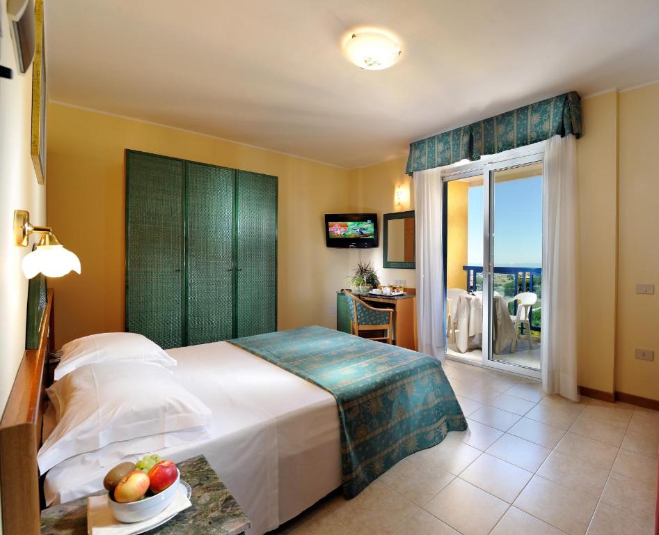 Mūsdienīga viesnīcas istaba ar divguļamo gultu, televizoru un balkonu, no kura paveras skats uz apkārtni. Uz galda ir augļu trauks, un istaba ir gaiša un ērta.