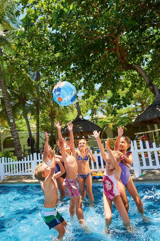 Bērni spēlējas baseinā, lēkājot un mēģinot noķert bumbu, kamēr apkārt ir zaļi koki un saulaina laika.