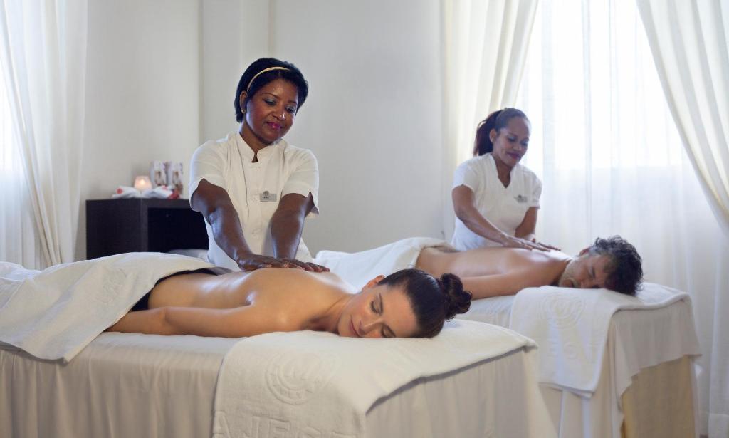 Relaksējoša masāža spa centrā, kur divi masāžas terapeiti strādā pie klientiem uz masāžas galdiem. Interjers ir gaišs un mierīgs, radot relaksējošu atmosfēru.