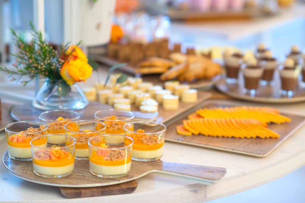 Krāšņi deserti, tostarp krēmveida pudiņi ar augļu dekoriem un sagriezti augļi, izkārtoti uz elegantas galda virsmas. Fons rotā ziedi un dažādi konditorejas izstrādājumi.