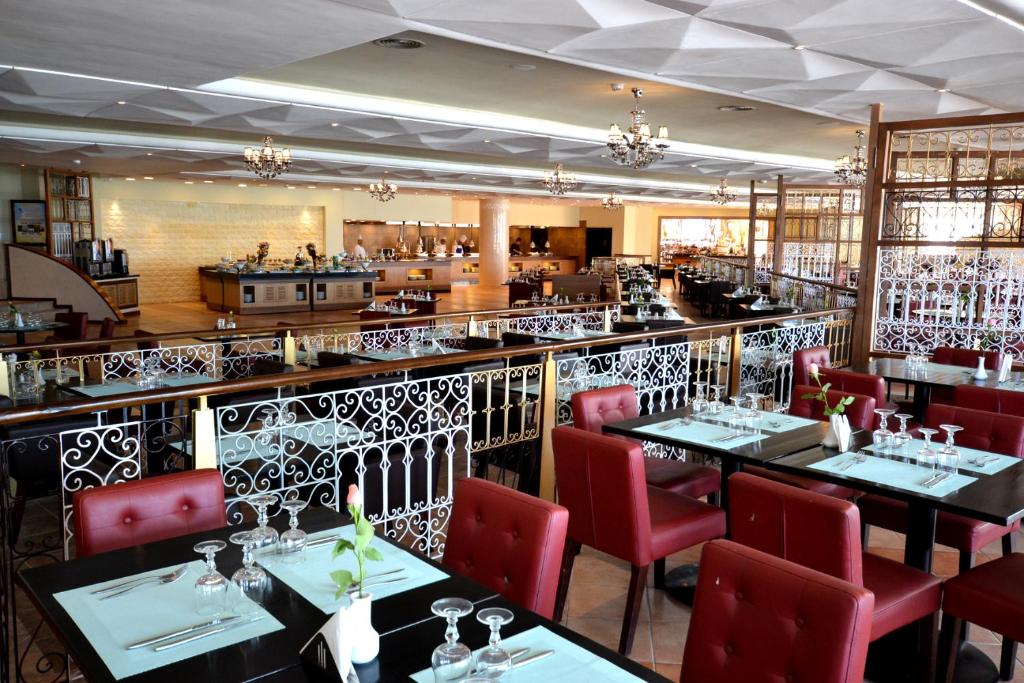 Elegants restorāna interjers ar sarkano krēslu un galdu izkārtojumu, dekorēts ar smalkām detaļām un kristāla lustru. Gaisotne ir stilīga un aicinoša, piemērota gan vakariņām, gan svinībām.