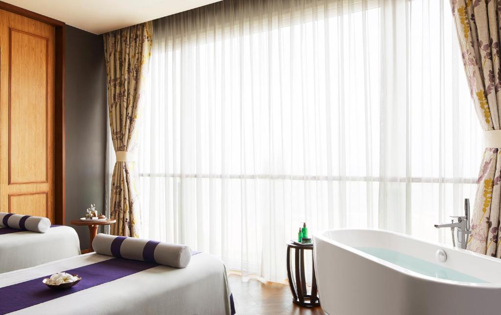 Spa telpa ar masāžas galdiem, elegantiem aizkariem un vannu, kas piedāvā relaksējošu atmosfēru. Gaisma plūst caur lielajiem logiem, radot mierīgu noskaņu.
