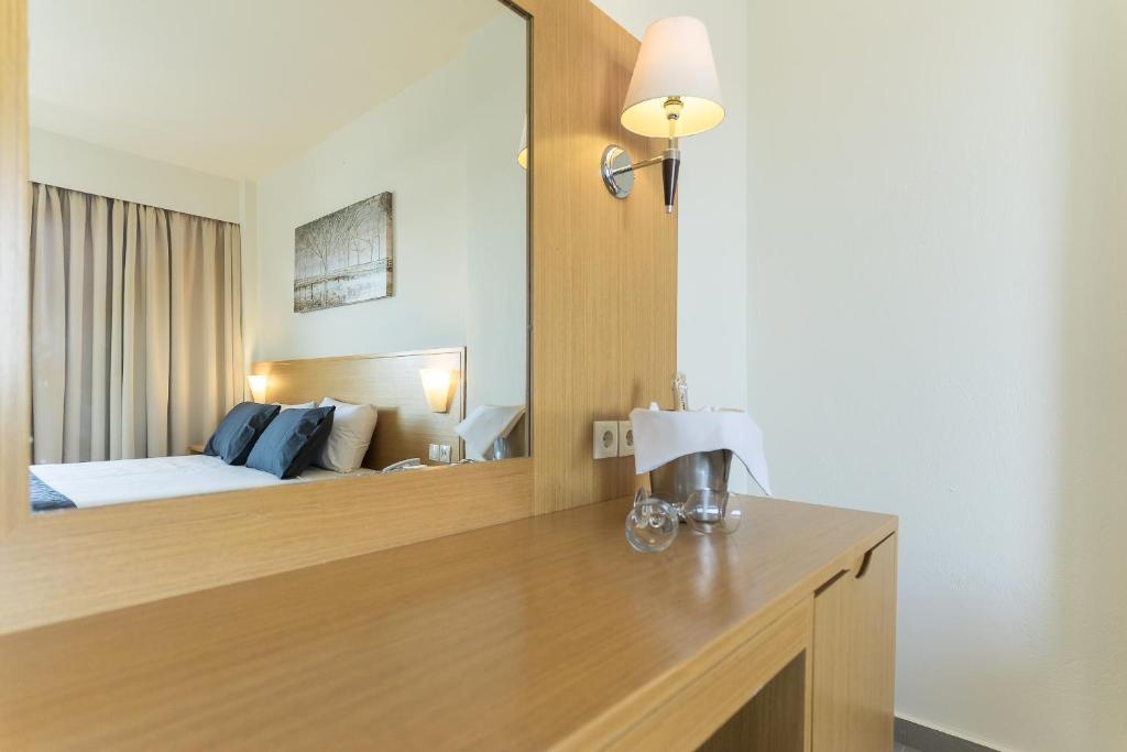 Mūsdienīga viesnīcas istaba ar koka mēbelēm, gultu un dekoratīvām lampām, atspoguļota spogulī. Uz galda atrodas glāzes un ledus spainis.