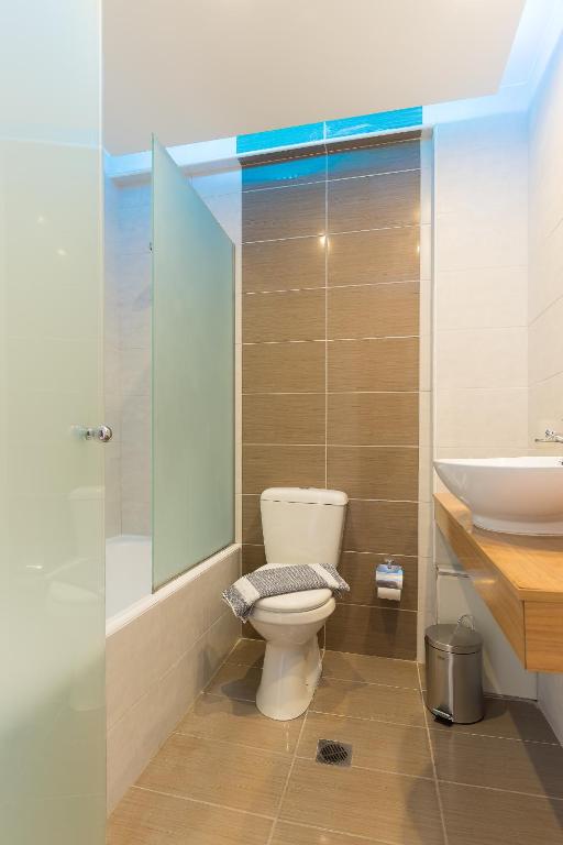 Mūsdienīgs vannas istabas interjers ar stikla dušu, tualeti un koka izlietni. Sienas ir apšūtas ar gaišiem flīzēm, radot elegantu atmosfēru.