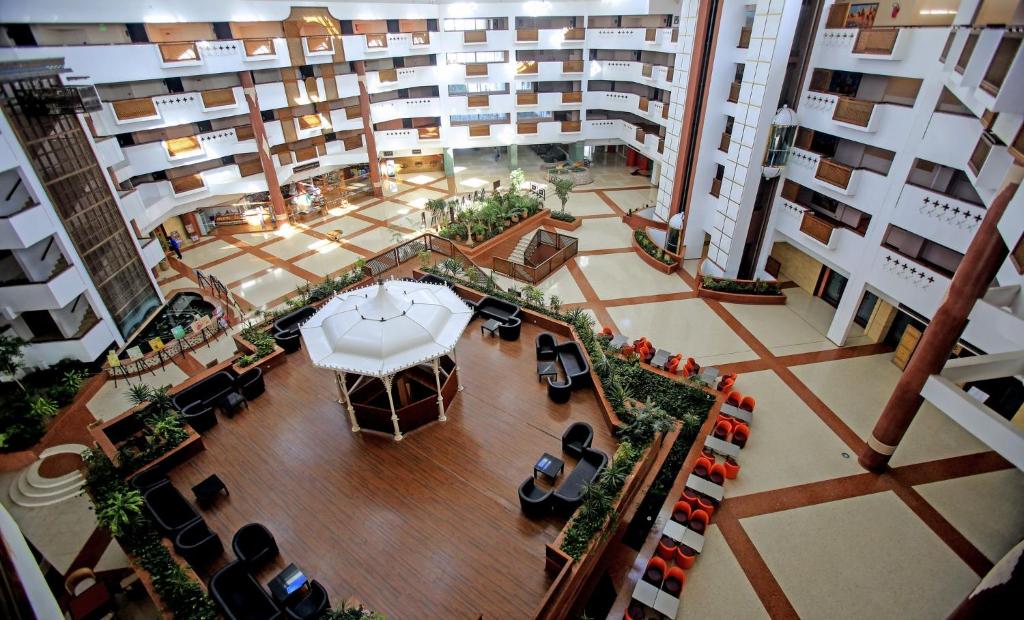 Mūsdienīga viesnīcas iekštelpa ar plašu atvērtu telpu, centrālais paviljons un komfortabli sēdvietas apkārtējā dārzā. Viegli redzama augsta arhitektūra un zaļumi.