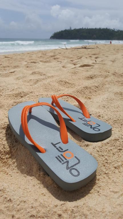 Sandales ar oranžiem siksniņām uz pludmales smiltīm, fons ar jūras viļņiem un zaļiem kalniem. Ideāla vasaras atmosfēra.