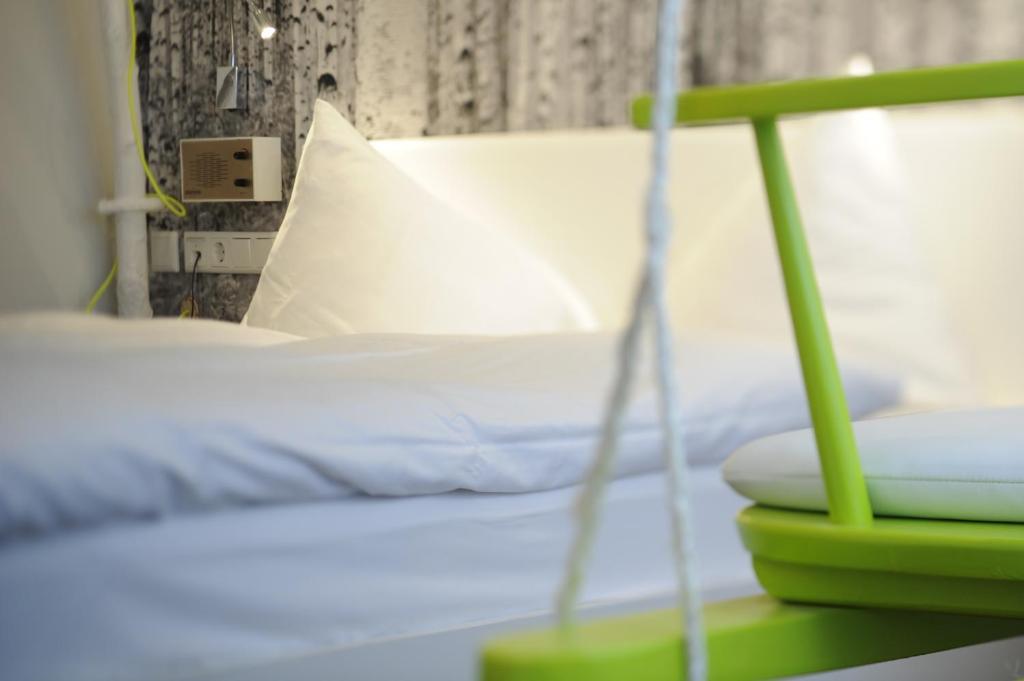 Mūsdienīgs guļamistabas interjers ar baltiem spilveniem un dīvānu, kas akcentēts ar zaļu šūpoļu dizainu. Fons ar koka tekstūru piešķir telpai siltumu un mājīgumu.