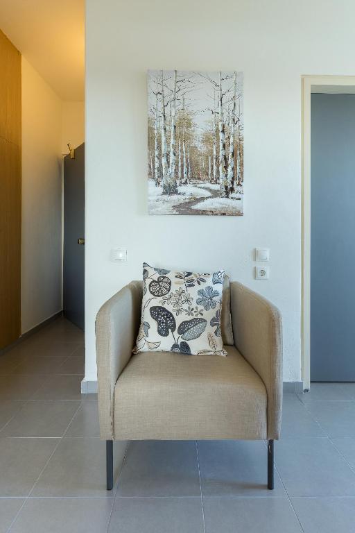 Mūsdienīgs krēsls ar dekoratīvu spilvenu, kas novietots gaišā telpā, uz sienas redzama ainava ar kokiem.