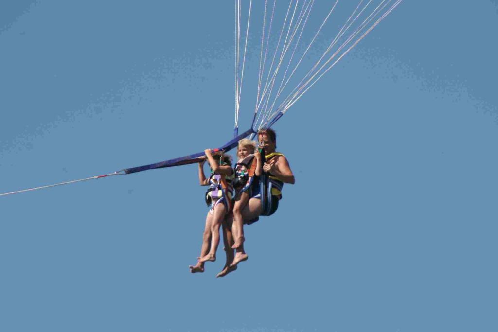 Divi cilvēki, kas piedalās parasailing, lido augstu debesīs ar krāsainiem izpletņiem. Skats ir skaidrs un saulains, radot piedzīvojuma sajūtu.