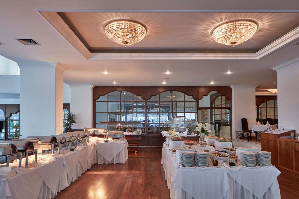 Elegantā restorāna iekārtojums ar bufetes galdu, kas klāts ar dažādiem ēdieniem un dekoratīvām ziedu kompozīcijām. Gaismas ķermeņi piešķir telpai siltu atmosfēru.