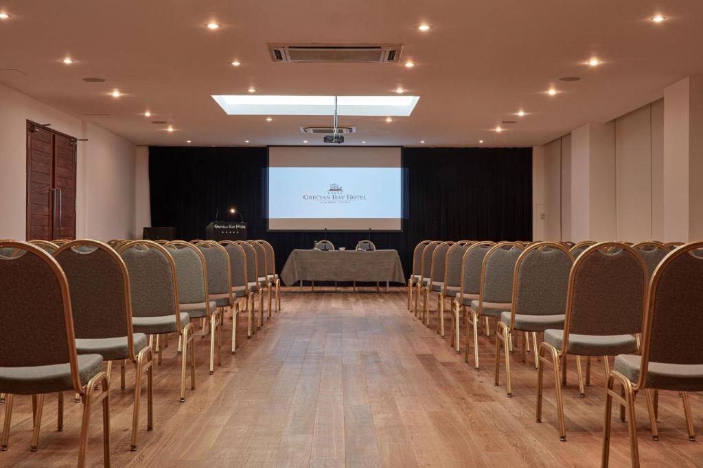 Konferenču telpa Grecian Bay Hotel ar rindu krēsliem un projekcijas ekrānu priekšā, radot profesionālu vidi sanāksmēm un prezentācijām.