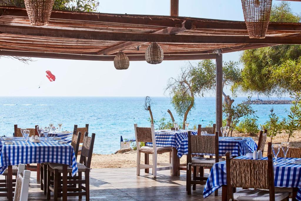 Vasaras restorāns ar zilu un baltu pārklāju galdiem, kas atrodas pie jūras. Skats uz okeānu un parasailing aktivitāte fonā.