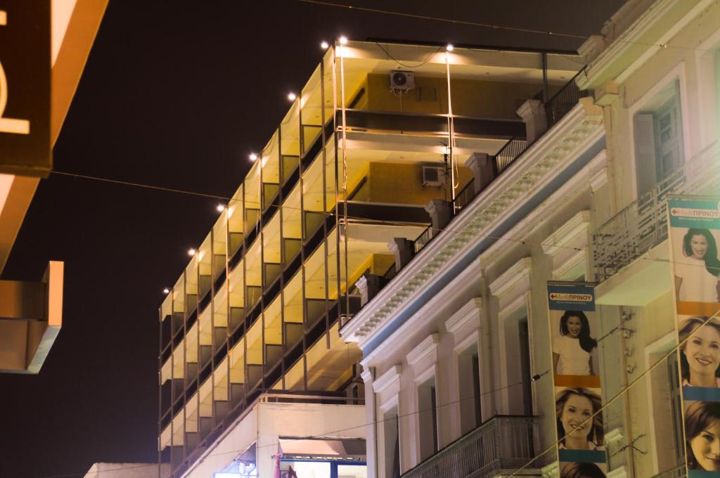 Naktī izgaismots ēku komplekss ar balkoniem un blakus esoša vēsturiska ēka, kurā redzami reklāmas baneri ar cilvēku attēliem.