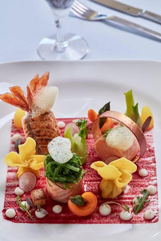 Mūsdienīgs gastronomijas mākslas darbs, kas ietver garneles, krāsainus dārzeņus un izsmalcinātas dekorācijas uz plāksnes ar mērci.