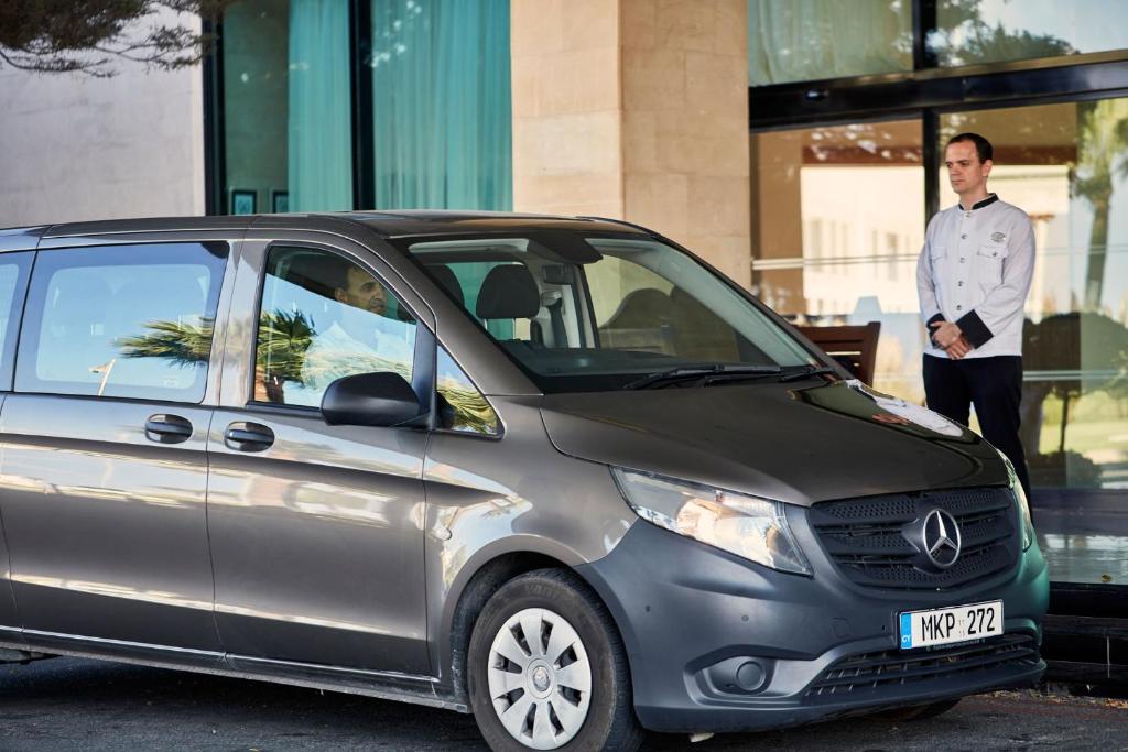 Melns Mercedes-Benz mikroautobuss stāv pie viesnīcas ieejas, kamēr viesmīlis gaida viesus. Fonā redzamas palmas un viesnīcas eleganta arhitektūra.