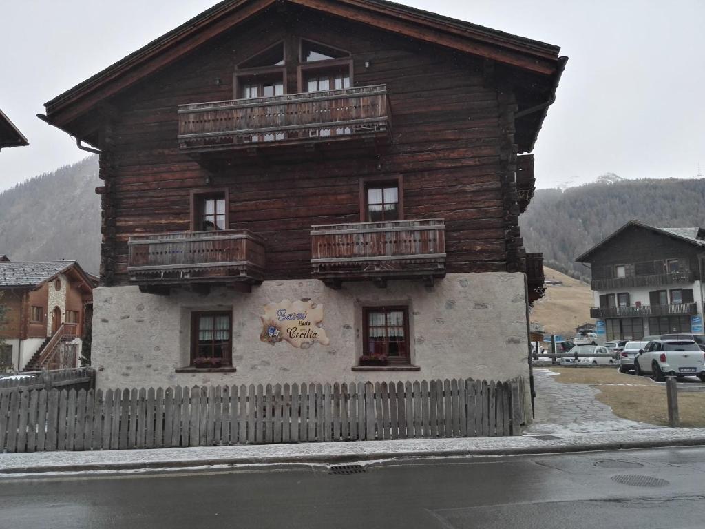 Tradicionāla kalnu māja ar koka fasādi un balkoniem, kas atrodas gleznainā ainavā. Priekšā redzams žogs un apkārtējā teritorija ar automašīnām.