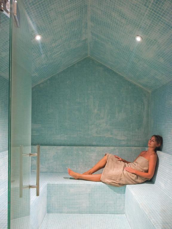 Mūsdienīga tvaika pirts ar gaiši zilu mozaīkas apdari, kurā atpūšas sieviete. Relaksējoša atmosfēra un mierīgs dizains.