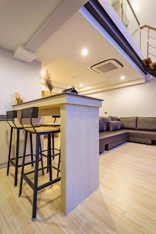 Moderns interjers ar bāra galdu un krēsliem, kas izvietoti atvērtā telpā, kurā redzama dīvāna zona. Siltas gaismas un koka grīdas piešķir mājīgu atmosfēru.