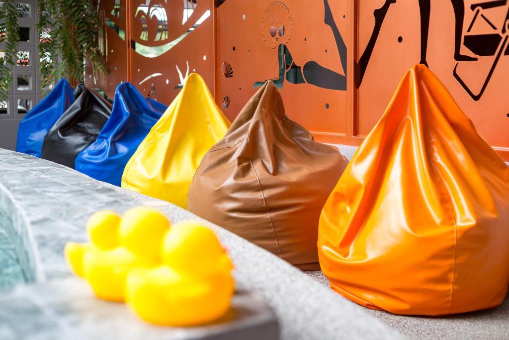 Krāsainu pupiņu maisu grupa, kas izvietota pie sienas ar radošu grafiku, radot jautru un relaksējošu atmosfēru. Priekšplānā redzami dzelteni gumijas pīlēni.