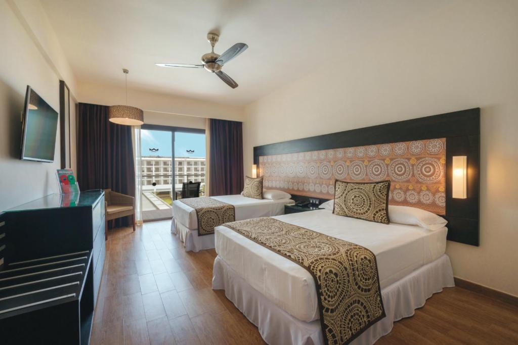 Mūsdienīga viesnīcas numura iekārtojums ar divām gultām, televizoru un balkonu, kas ved uz ārpusi. Telpai ir gaišas krāsas un dekoratīvi elementi uz sienas, radot patīkamu atmosfēru.